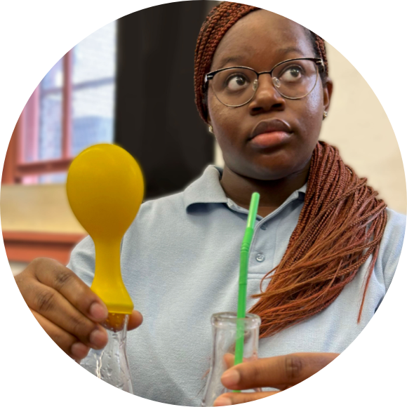 A person in glasses holds a yellow balloon over a glass container with liquid, while also holding a green straw.