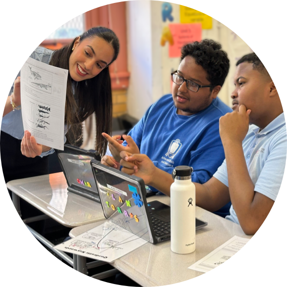 A teacher stands by two students seated at desks, showing them a paper while they work on their laptops. The students are engaged and focused, with one holding a pen and the other wearing glasses.