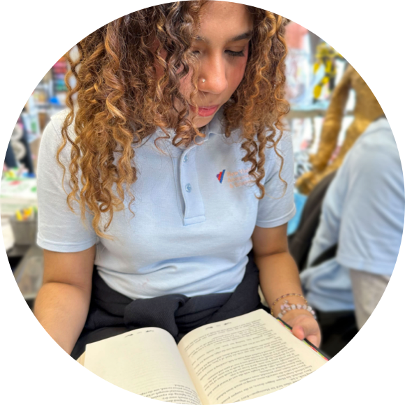 Young woman with curly hair reading a book while wearing a light blue polo shirt, seated in what appears to be a casual setting.