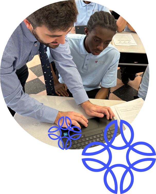 An instructor in a striped shirt assists a student with a braided hairstyle on a laptop at a table, while other students work in the background.