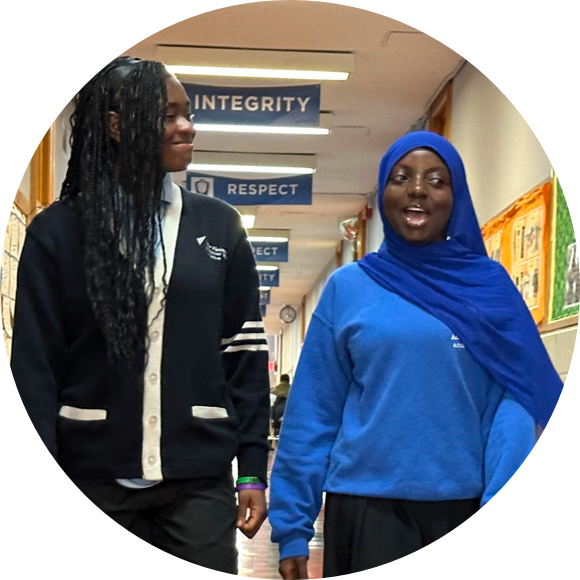 Two students in school uniforms walking down a hallway adorned with banners that read "INTEGRITY" and "RESPECT." One wears a cardigan and the other a blue hijab.