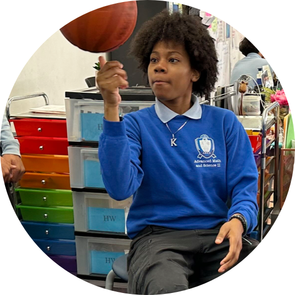A person in a blue sweatshirt spins a basketball on their finger while sitting in a classroom with colorful drawers in the background.