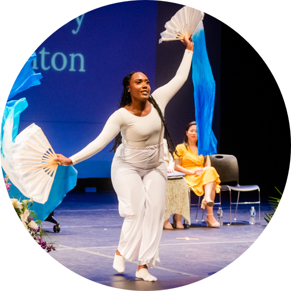 A performer wearing a white outfit dances with two white fans on stage, while another person in a yellow dress sits in the background.