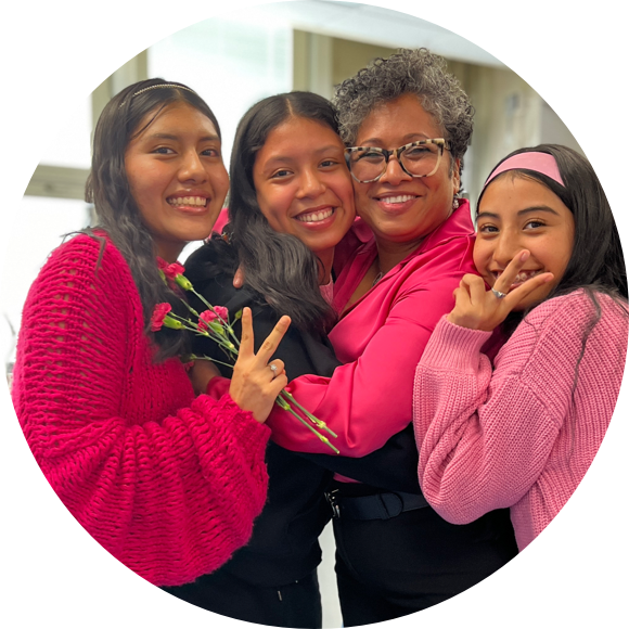 Four people, three young women and one older woman, smiling and hugging. The younger women hold flowers and wear pink tops, while the older woman wears glasses and a pink blouse.