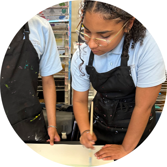 Two individuals wearing black aprons and light blue shirts are painting. One person is focused on painting while the other points at something on the table.