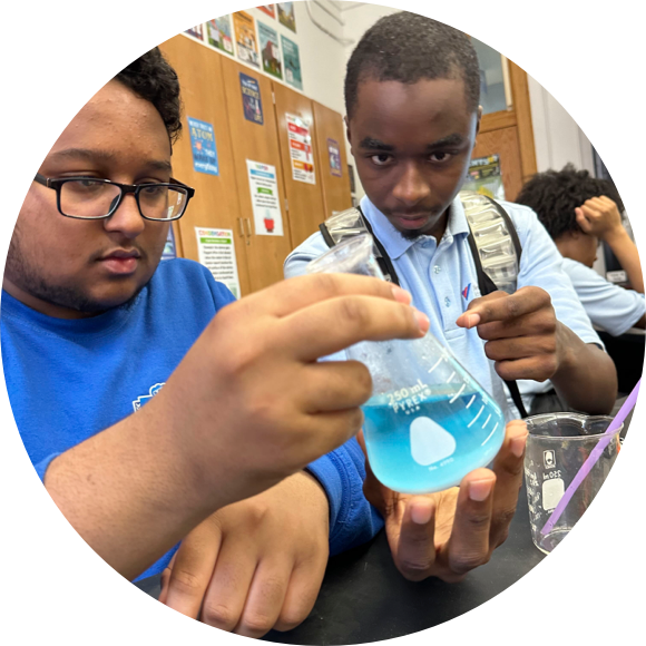 Two students in a classroom look intently at a beaker filled with blue liquid. One student is wearing glasses and a blue shirt, while the other has a serious expression and is holding the beaker.