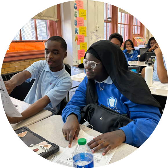 Two students in a classroom are looking at a paper and discussing it. Both are seated at desks with worksheets and water bottles in front of them. Other students are visible in the background.