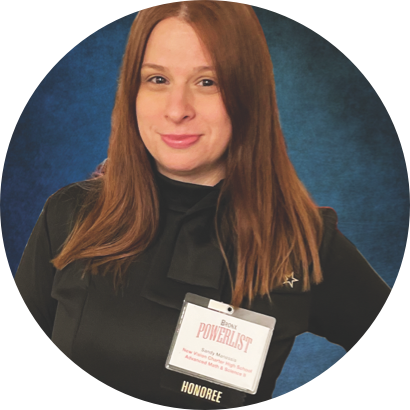 A woman with long red hair is wearing a black top and a name badge labeled "Bronx Powerlist Honoree" against a blue background.