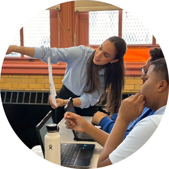 A woman in a light blue shirt explains a document to two seated individuals using laptops in a classroom setting.