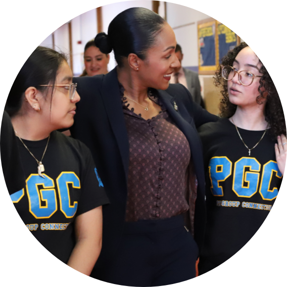 A woman in business attire stands with two young people in matching PGC t-shirts, gesturing and engaging in conversation.