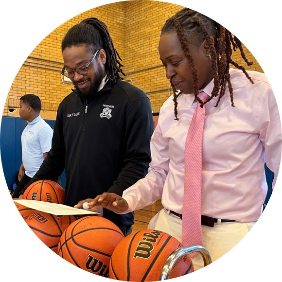 Two people stand near a rack of basketballs in a gym, looking at a document. One wears a black jacket and the other a pink shirt and tie. A third person is in the background.