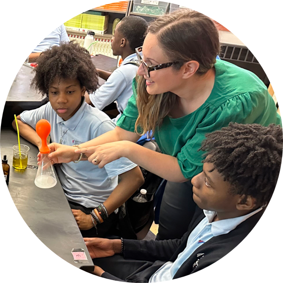 A teacher in a green shirt demonstrates a science experiment to two attentive students. A balloon inflates over a beaker containing white foam.