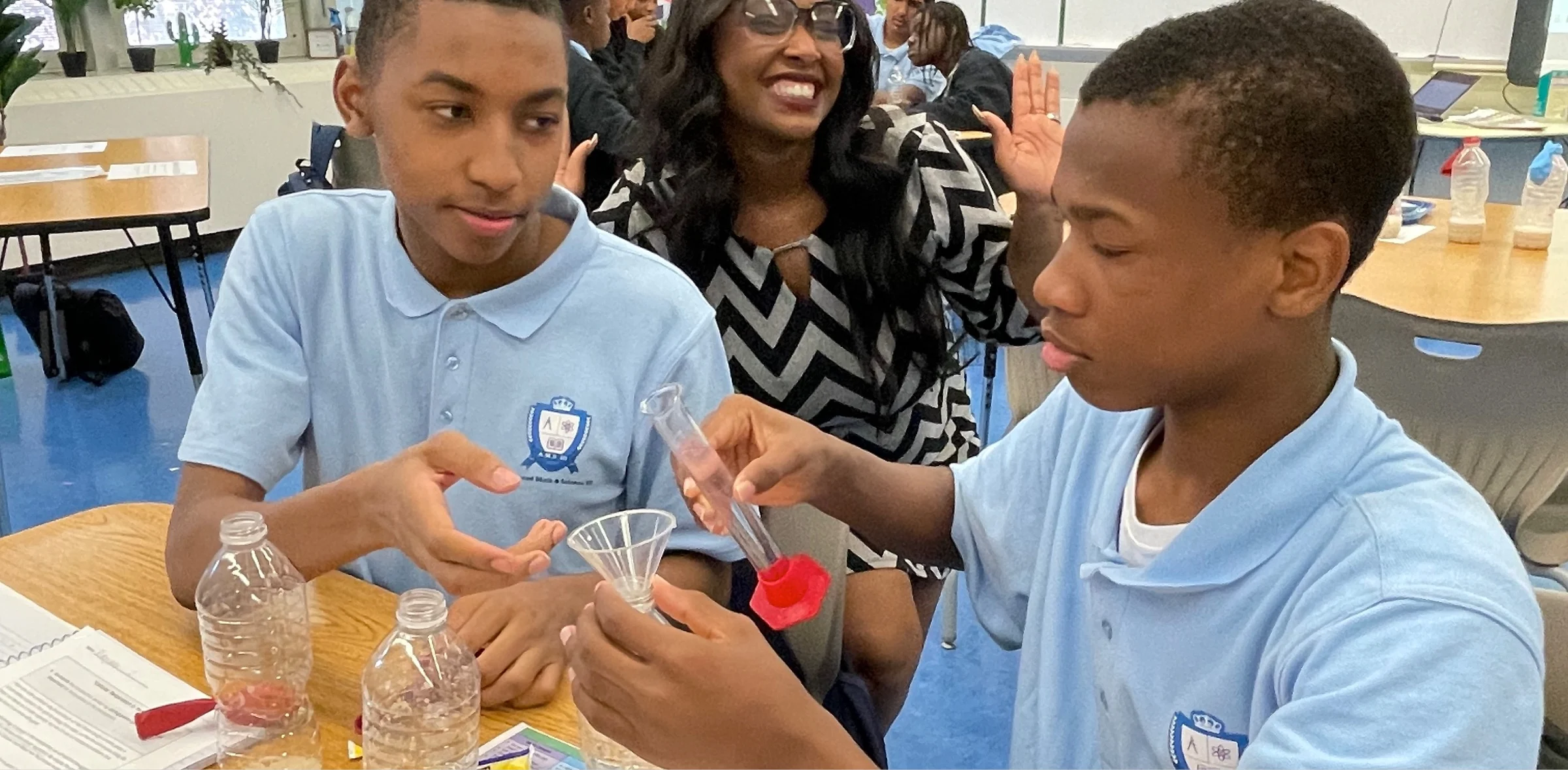 Two students in blue uniforms conduct a science experiment with a funnel and test tube at a table, while a teacher smiles in the background.
