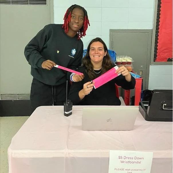Two people at a table with a laptop, selling pink wristbands for $5. A sign reads, "$5 Dress Down Wristbands!" They are smiling and standing indoors.