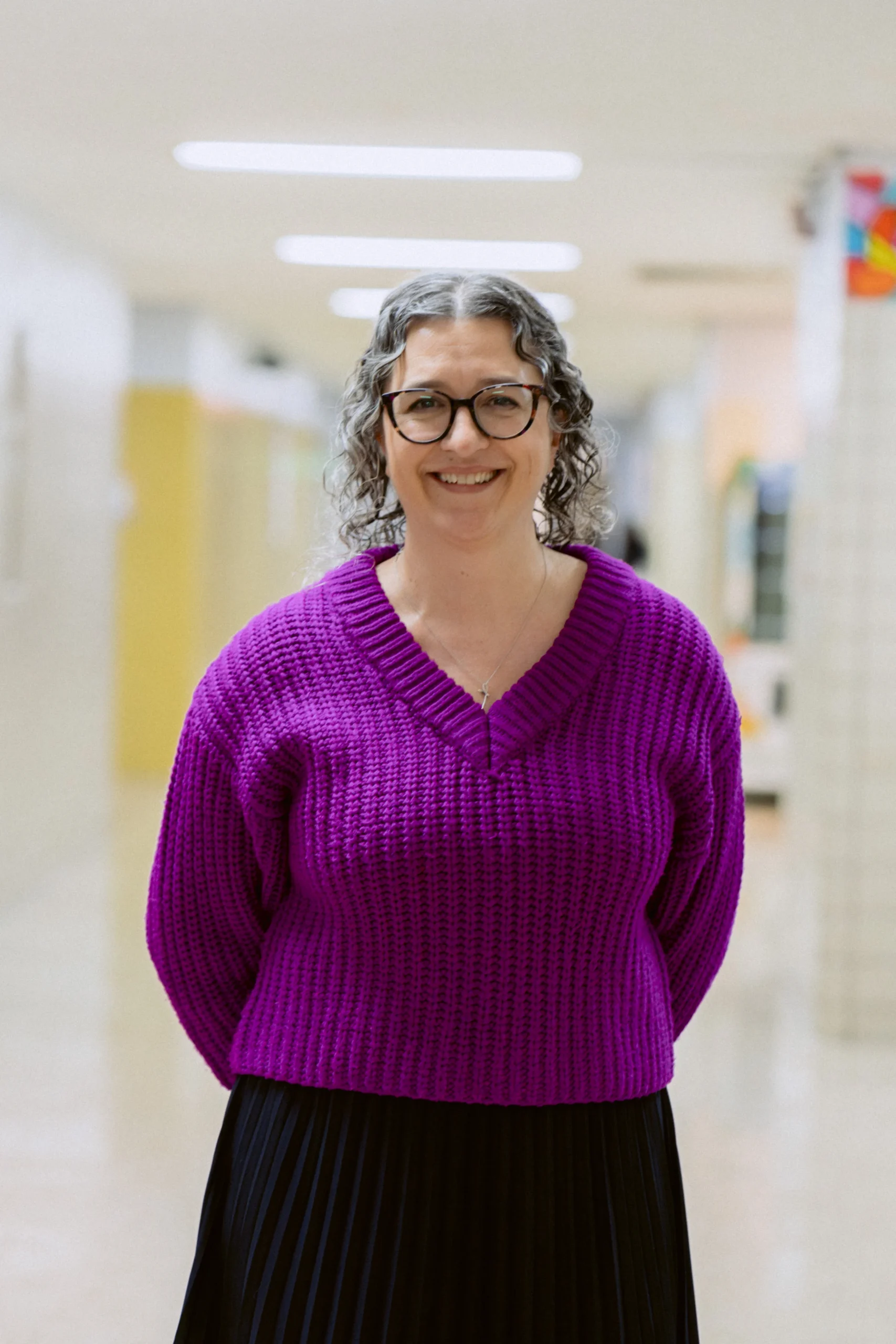 A woman with curly gray hair and glasses smiles, wearing a purple sweater and black skirt, standing in a brightly lit hallway.