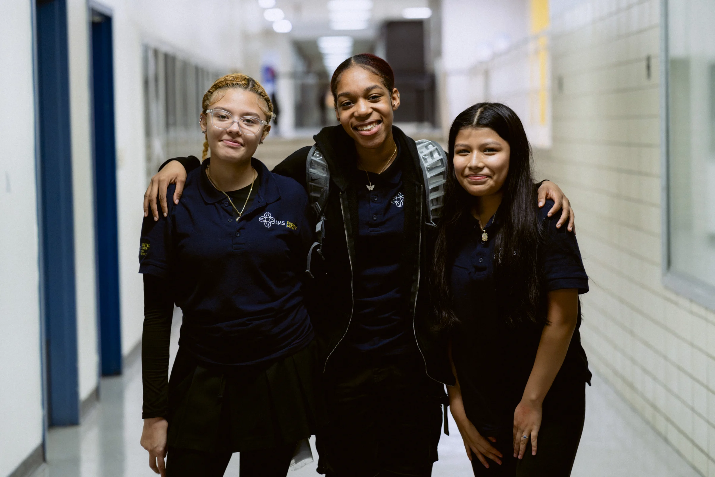 Three students in school uniforms stand in a hallway, smiling and posing for the camera.