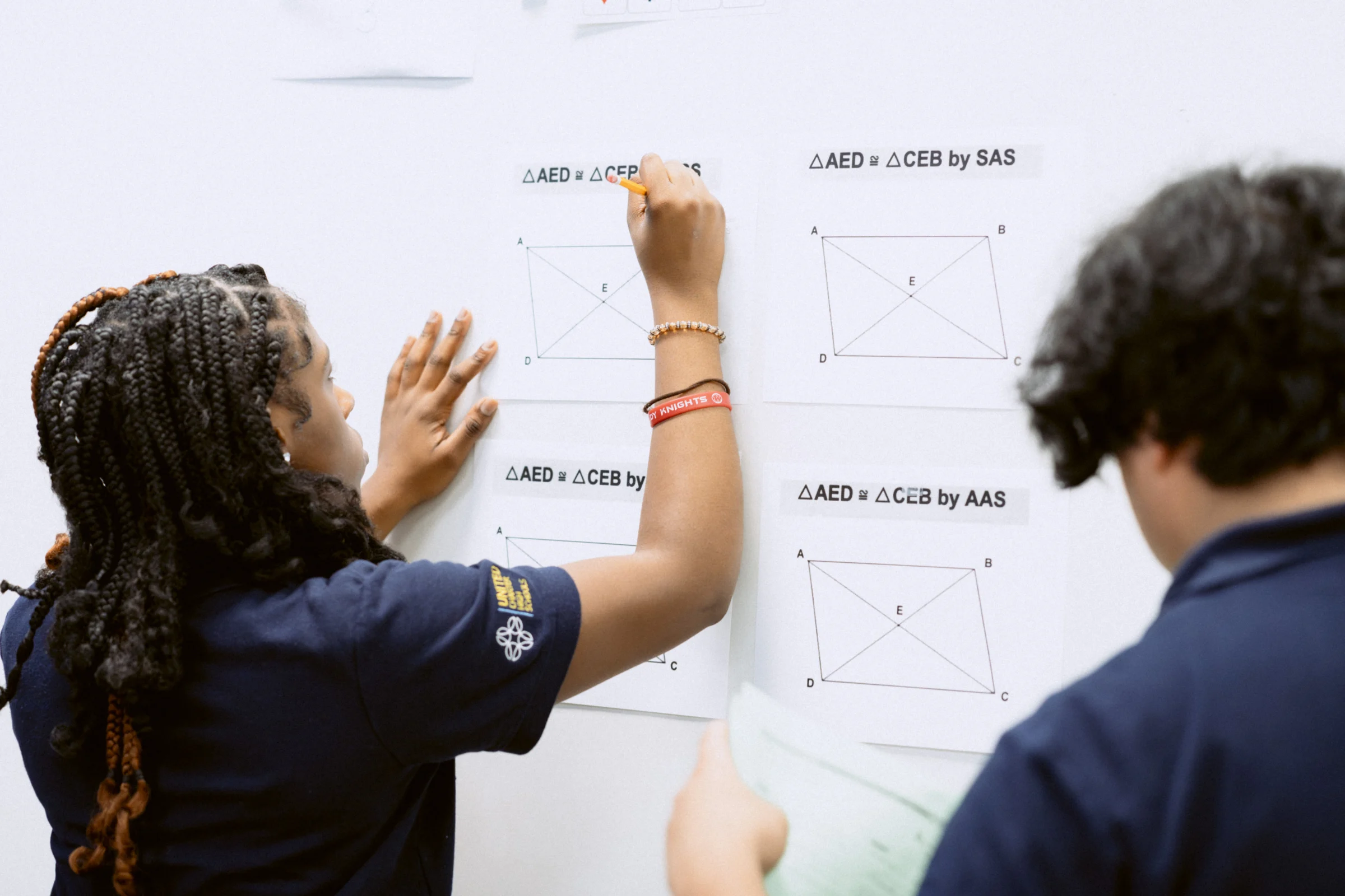 Two students in dark blue shirts solve geometry problems on a whiteboard, using diagrams and symbols to demonstrate triangle congruency theorems like SAS and AAS.