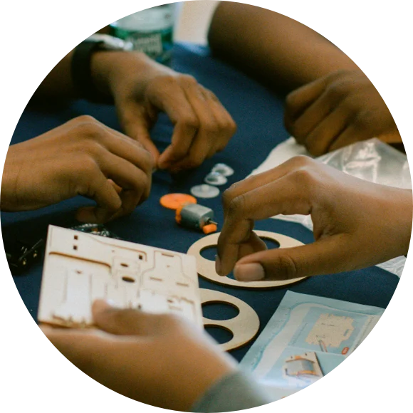 Hands assembling a model with wooden pieces, small components, and a small motor on a blue table.
