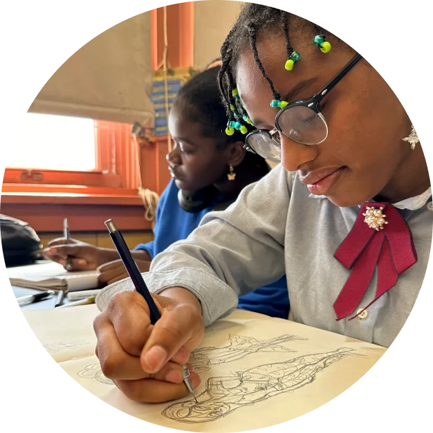 Two students sit at desks, focused on sketching in notebooks. One wears glasses and has colorful beads in hair. Bright window in the background.