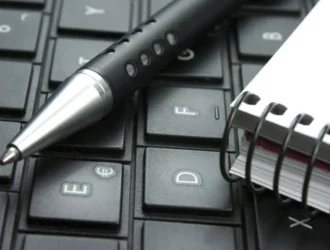 A pen and a spiral notebook are placed on a black computer keyboard.