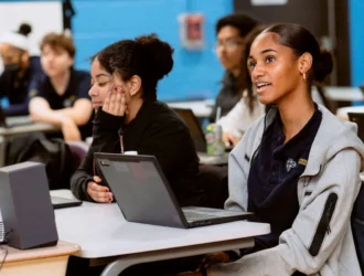 A student at United Charter High Schools sits at a desk in a classroom and looks up. There are students at tables and each one has a computer.