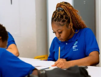 Student in blue school uniform focuses on writing at a desk, surrounded by classmates in a classroom setting.