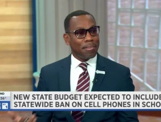 A man in a suit and glasses speaks on a news program. The screen headline discusses a possible statewide ban on cell phones in schools.