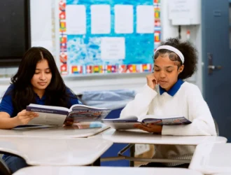 Two students United Charter High Schools HUM sit at desks in a classroom, reading textbooks. One wears a blue shirt and the other a white sweater. Classroom materials and bulletin board are visible in the background.