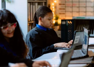 Three United High Schools students sit at desks using laptops in a classroom with a whiteboard and shelves in the background. The focus is on the student in the center.