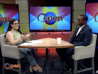 A woman interviews a man in a studio set with "Open" displayed on monitors behind them; both are seated at a table with papers and a cup.