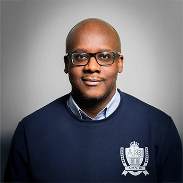 A man with glasses wearing a navy sweater with a crest emblem, posing against a plain grey background.