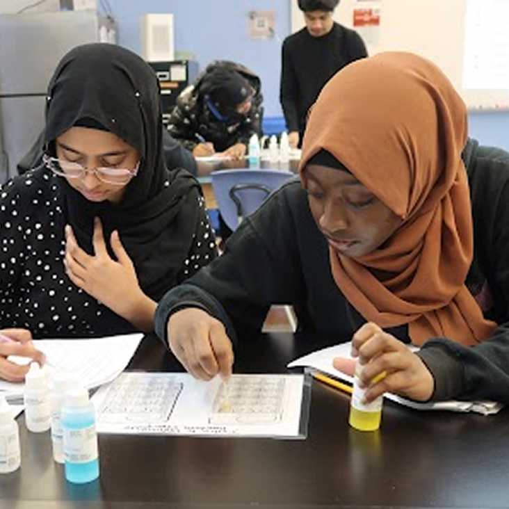 Two students wearing hijabs work together at a classroom desk with worksheets and bottles, while others work in the background.