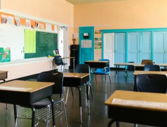 Empty classroom with desks arranged in rows, a green chalkboard on one wall, and blue doors and cabinets at the back.