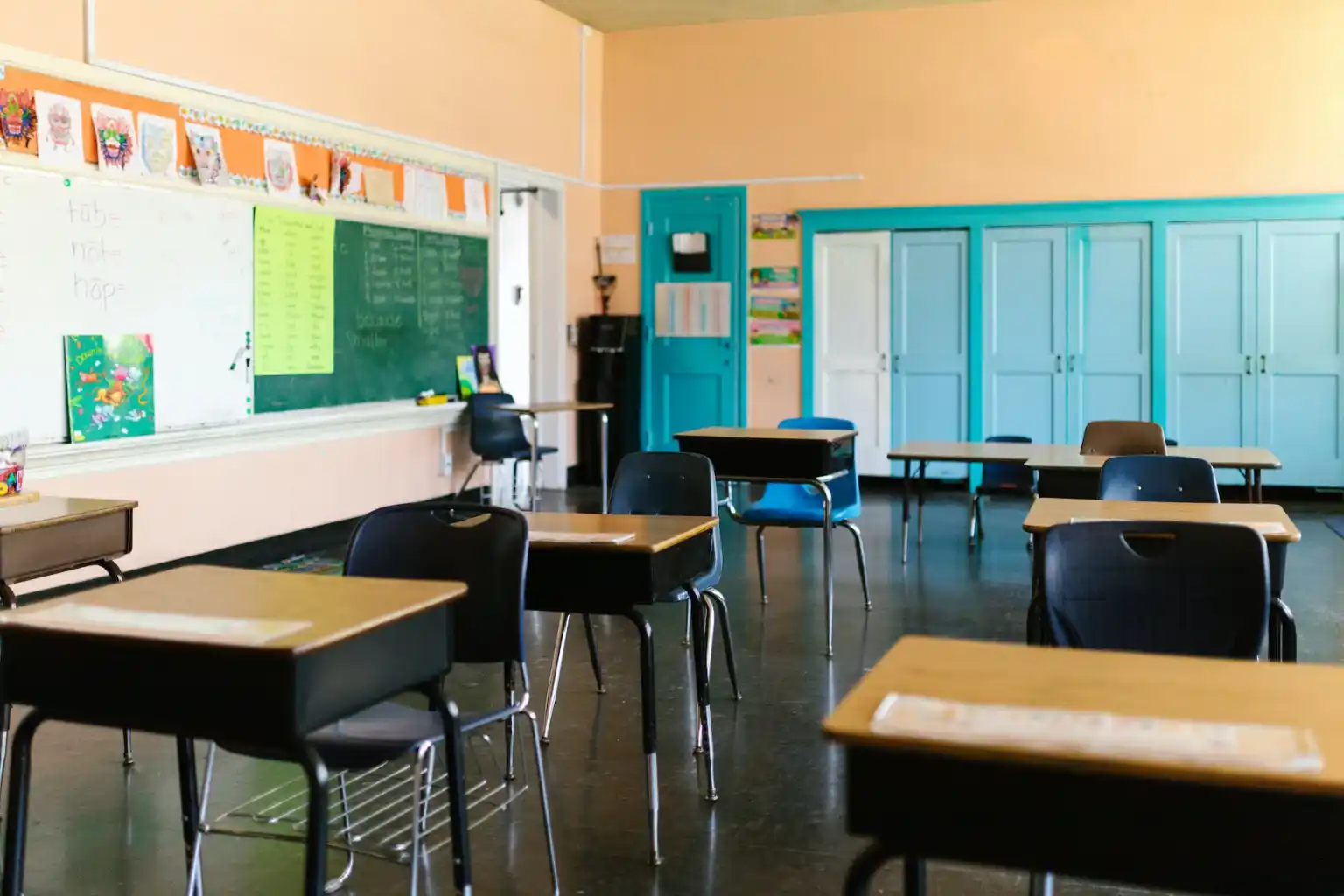 Empty classroom with desks arranged in rows, a green chalkboard on one wall, and blue doors and cabinets at the back.