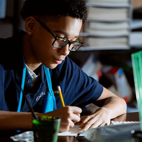 A young person wearing glasses and a blue shirt writes on paper at a desk, with art supplies visible nearby.