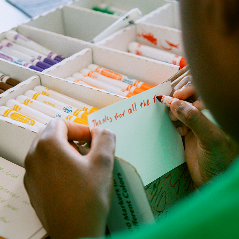 A person writes "Thanks for all the" in red marker on a card, with boxes of colored markers visible in the background.