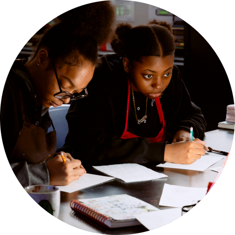 Two young people sit at a table, focused on writing or drawing on sheets of paper, with notebooks and supplies nearby.