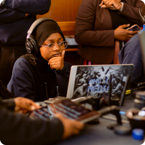 A person wearing a headset sits in front of a laptop and audio equipment, appearing focused while working in a media production setting.