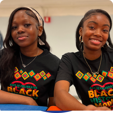 Two young women sit side by side, smiling, both wearing matching "Black History Month" t-shirts with colorful designs.
