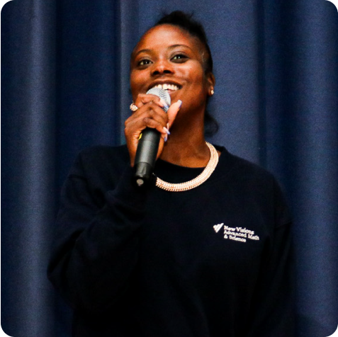 A person stands in front of a blue curtain, holding a microphone and smiling, wearing a dark sweatshirt with a logo on the left side.