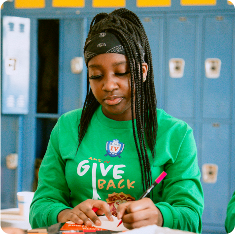 A young person in a green "Give Back" shirt writes with a pink marker at a table, with blue lockers in the background.