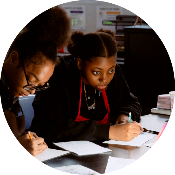 Two students sit at a desk, focused on writing or drawing on paper with pencils, in a classroom setting.