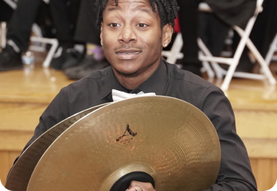 A person dressed in formal black attire holds a cymbal during a musical performance on a stage with chairs in the background.