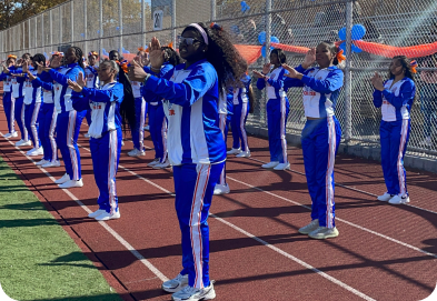 A group of cheerleaders in blue and white uniforms perform a routine on a running track beside a chain-link fence decorated with balloons.